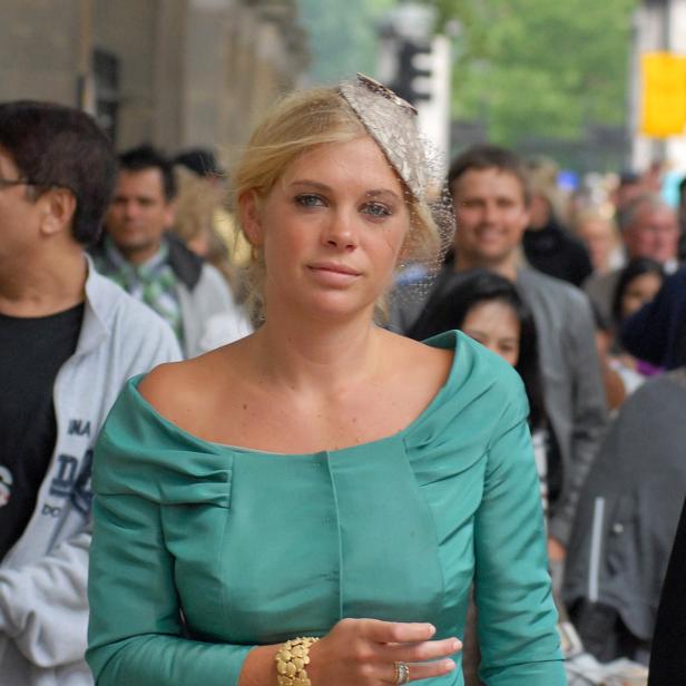 Eine Frau in einem türkisfarbenen Kostüm und einem Fascinator geht durch eine Menschenmenge.
