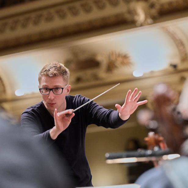 Ein Dirigent mit Brille dirigiert ein Orchester.
