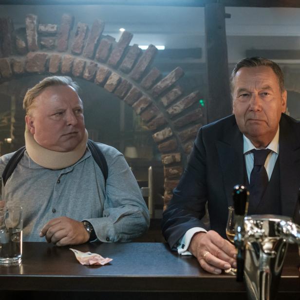 Zwei Männer sitzen an einer Bar, einer trägt eine Halskrause.