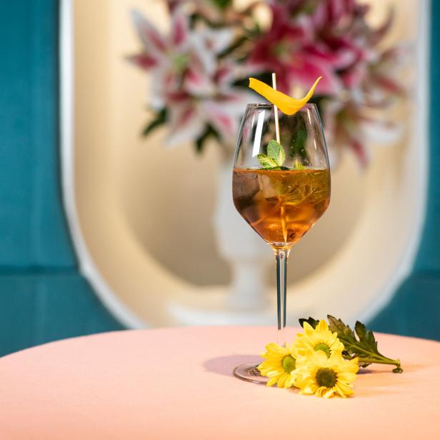 Ein Cocktail mit Minze und einer Orangenzeste steht auf einem Tisch mit gelben Blumen.