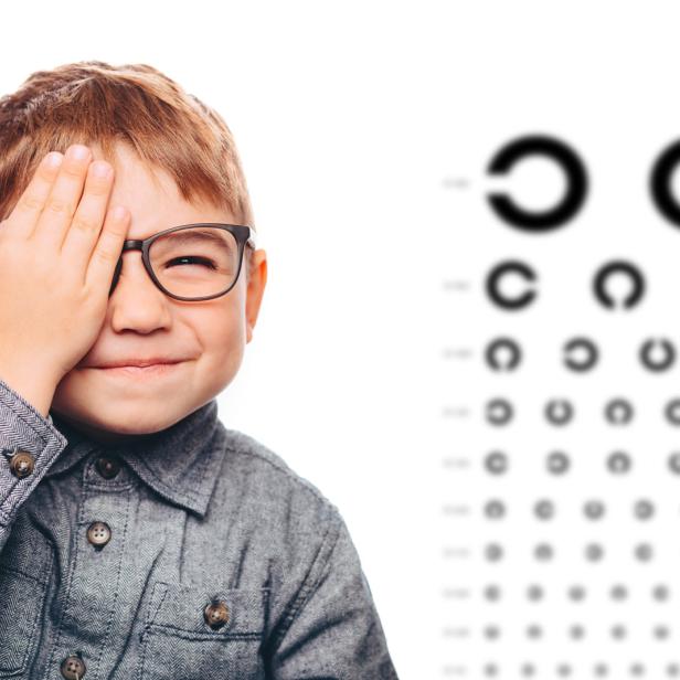 Ein Junge mit Brille hält eine Hand vor ein Auge, im Hintergrund eine Sehtafel.