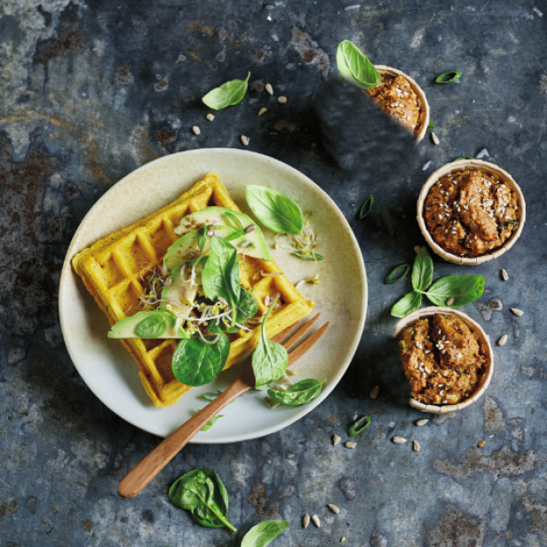 Ein herzhaftes Waffelgericht mit Avocado, Sprossen und Basilikum auf einem Teller.