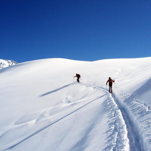 Zwei Skibergsteiger steigen einen verschneiten Hang unter blauem Himmel hinauf.