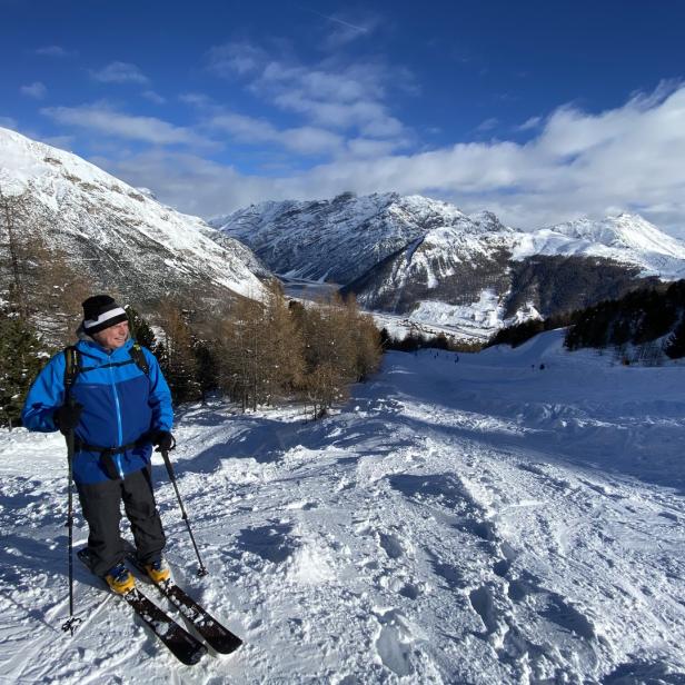 Ein Skifahrer steht auf einer Skipiste mit schneebedeckten Bergen im Hintergrund.