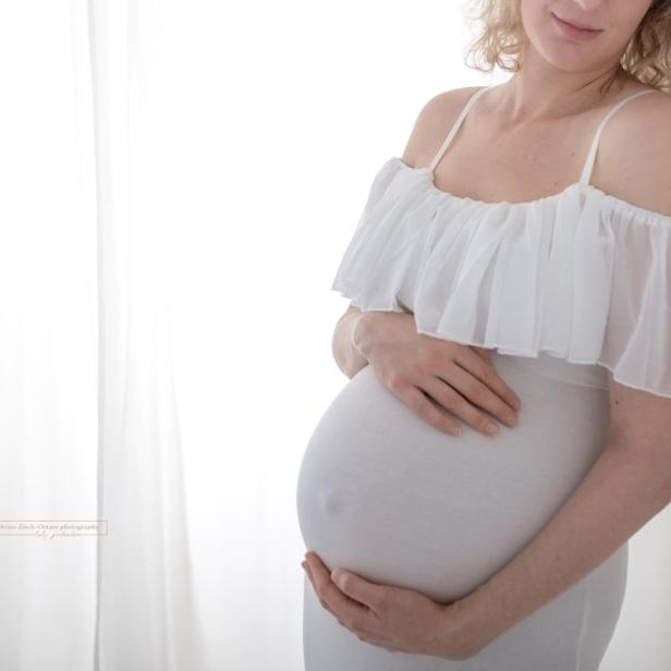 Eine schwangere Frau in einem weißen Kleid hält ihre Hände auf ihren Bauch.