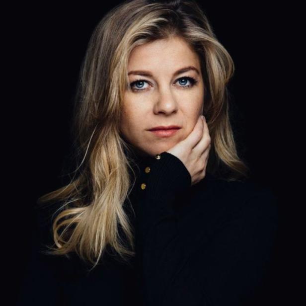 Eine Frau mit blonden Haaren und blauen Augen posiert vor einem schwarzen Hintergrund.