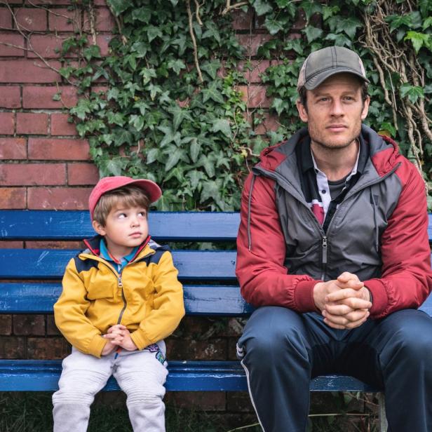 Ein Mann und ein kleiner Junge sitzen auf einer blauen Bank vor einer Ziegelmauer mit Efeu.