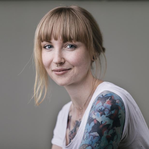 Eine Frau mit blonden Ponyfransen, Nasenring und Tätowierungen lächelt.