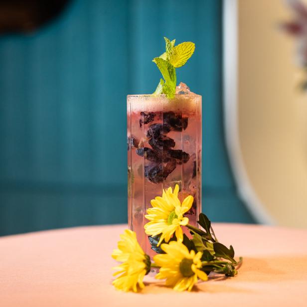 Ein Glas mit einem rosafarbenen Getränk, Blaubeeren und Minze garniert, steht auf einem Tisch.
