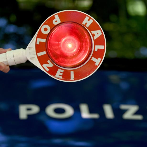 Die Hand eines Polizisten hält ein rotes Halte-Schild. Im Hintergrund ist ein Polizeiauto zu erkennen.