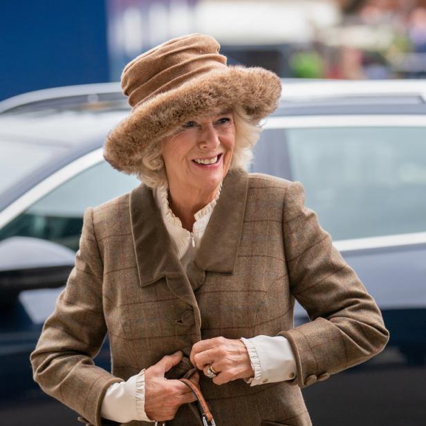 Königin Camilla lächelt mit Hut und braunem Tweed-Mantel und hält eine Handtasche.