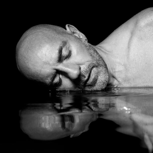 Ein Mann liegt mit geschlossenen Augen im Wasser, sein Spiegelbild ist sichtbar.