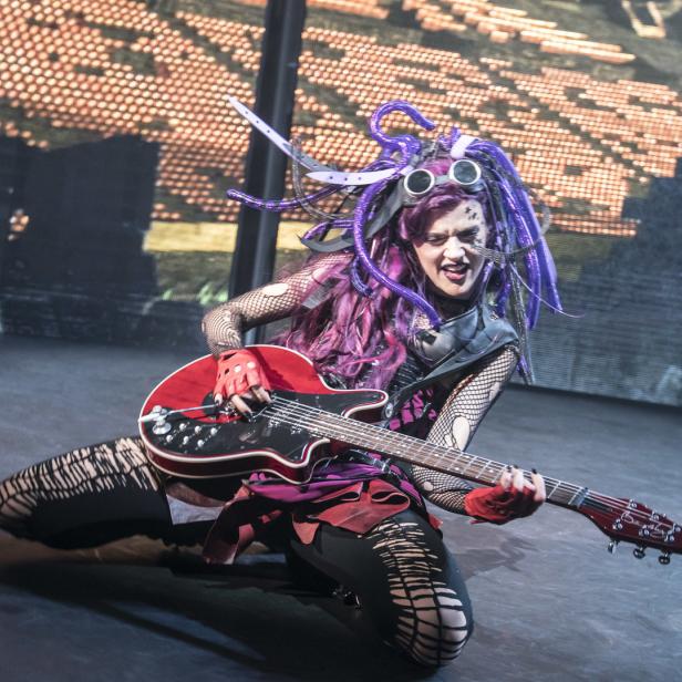 Eine Frau mit lila Haaren und futuristischer Kleidung spielt E-Gitarre auf einer Bühne.