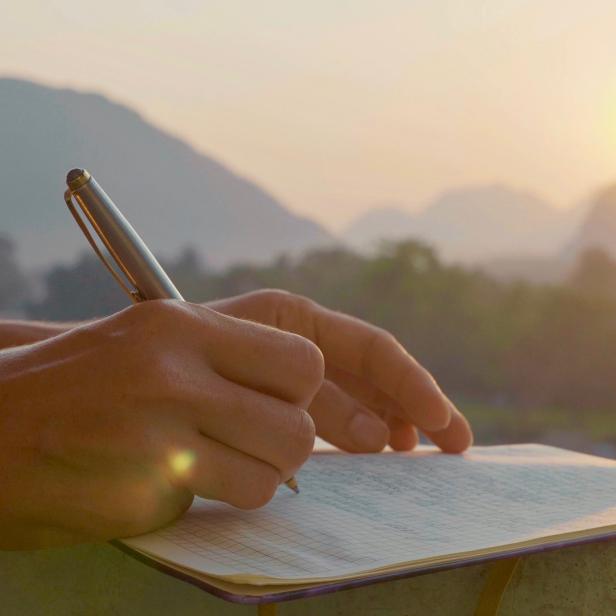 Eine Person schreibt mit einem Stift in ein Notizbuch vor einer Bergkulisse bei Sonnenaufgang.