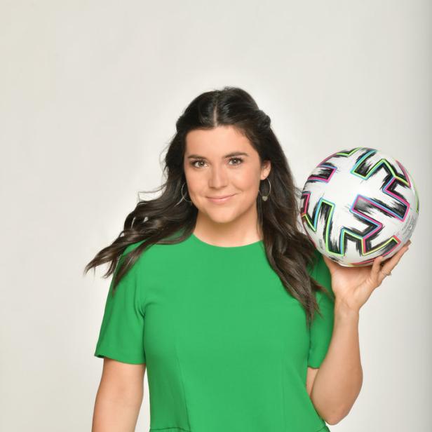 Eine Frau im grünen Oberteil hält einen Fußball in der Hand.