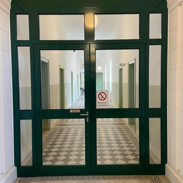 Eine grüne Doppeltür mit Glas führt in einen Flur mit Schachbrettmuster und einem "Rauchen verboten"-Schild.