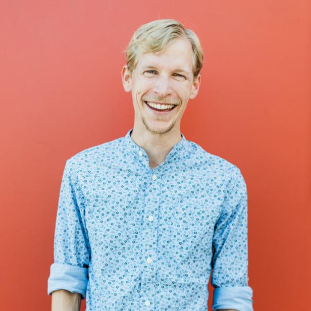 Ein lächelnder Mann mit blonden Haaren vor einer roten Wand.