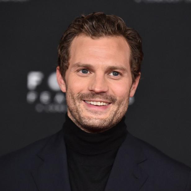 Porträt von Jamie Dornan, der in einem schwarzen Rollkragenpullover und einem dunklen Anzug lächelt.