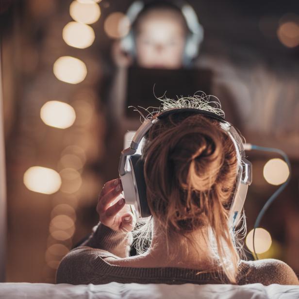 Eine junge Frau hört Musik über Kopfhörer, während eine weitere Person im Hintergrund ein Tablet benutzt.