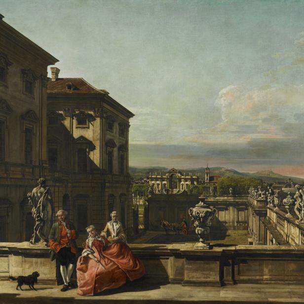 Ein Paar mit Hund auf einer Terrasse mit Blick auf ein Schloss und eine hügelige Landschaft.