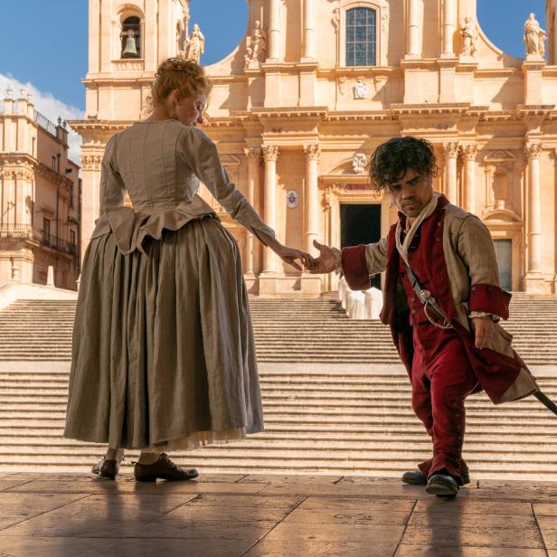 Peter Dinklage und eine Frau in historischer Kleidung stehen vor einem großen Gebäude.