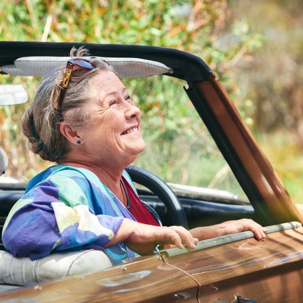 Eine ältere Frau lächelt, während sie in einem braunen Cabriolet fährt.