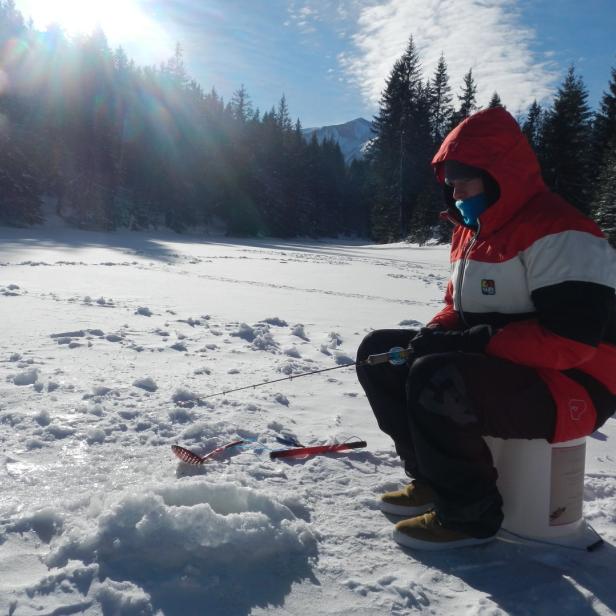 Ein Mann sitzt auf einem Eimer beim Eisangeln in einer verschneiten Landschaft.