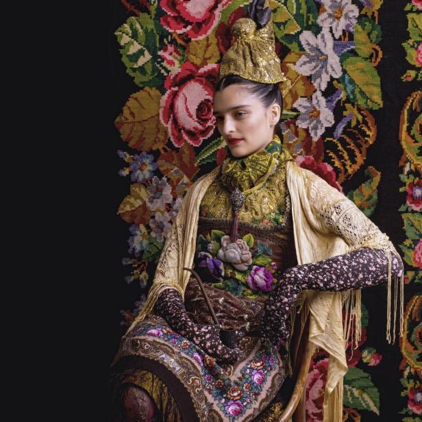 Eine Frau in traditioneller Tracht sitzt vor einem blumigen Wandteppich.