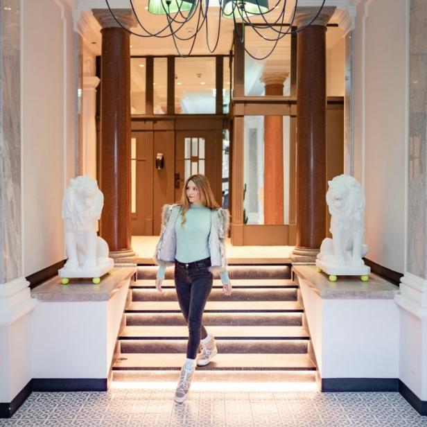 Eine Frau geht eine Treppe in einem eleganten Gebäude hinunter, unter einem Kronleuchter mit vielen Lampen.