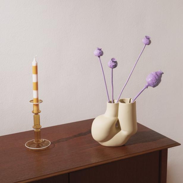 Eine gestreifte Kerze in einem goldenen Kerzenständer und lila Mohnblumen in einer beigen Vase auf einem Holzschrank.