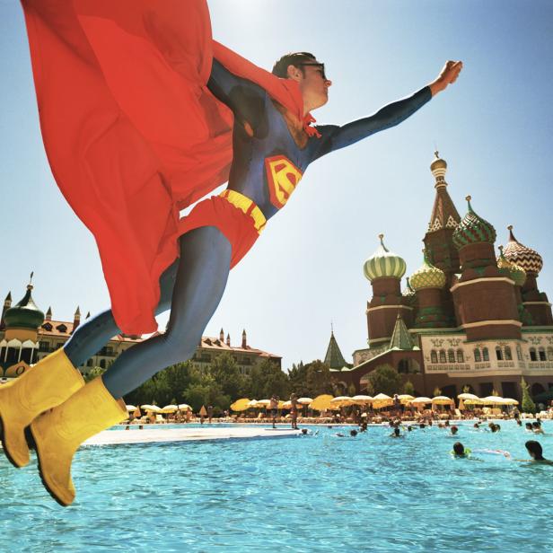 Ein Mann im Superman-Kostüm springt vor einem Schwimmbad mit russisch anmutender Architektur.