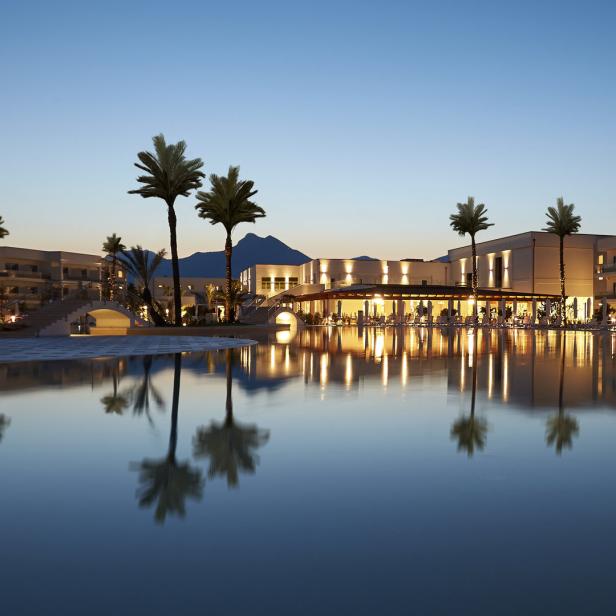 Ein Resort mit Palmen spiegelt sich im ruhigen Wasser eines Pools bei Dämmerung.
