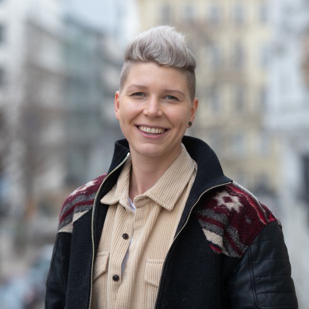 Eine lächelnde Frau mit kurzen, grauen Haaren und einer gemusterten Jacke steht im Freien.