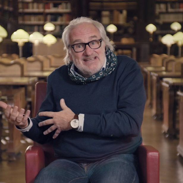 Ein Mann mit Brille gestikuliert in einer Bibliothek sitzend.