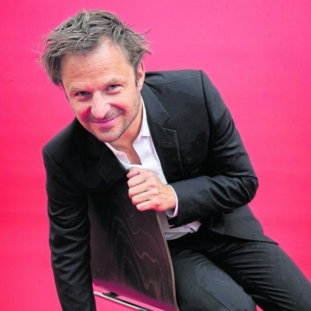 Philipp Hochmair sitzt auf einem Stuhl vor einem roten Hintergrund.