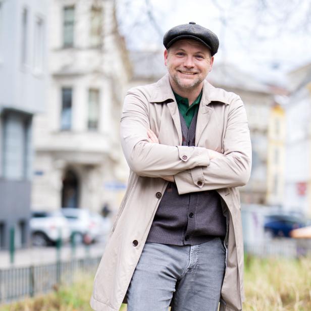 Ein lächelnder Mann mit Schirmmütze und Trenchcoat steht auf der Straße.