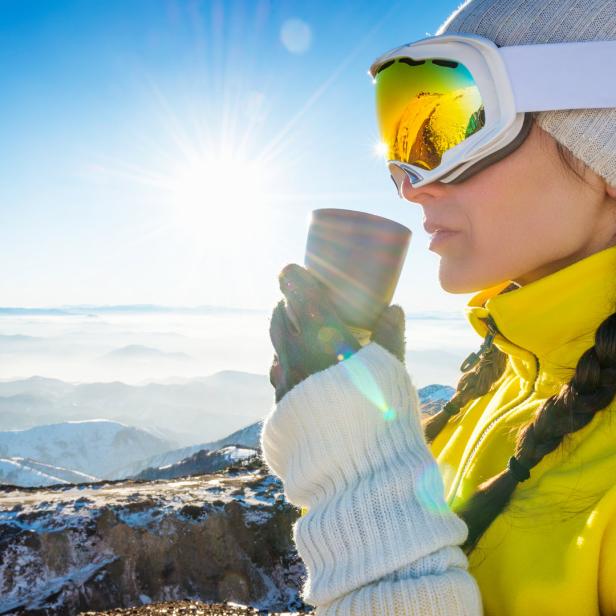 Eine Frau mit Skibrille trinkt auf einem schneebedeckten Berggipfel etwas Warmes.