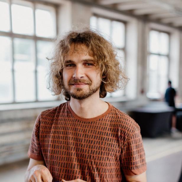 Ein Mann mit lockigem Haar hält einen Messbecher mit einer dunklen Flüssigkeit in der Hand.