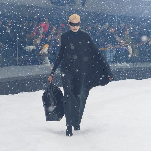 Eine Person in schwarzer Kleidung und Sonnenbrille läuft mit einer Mülltüte über einen verschneiten Laufsteg.