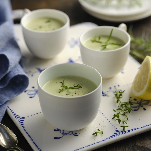 Drei Tassen cremige Suppe, garniert mit Kräutern, stehen auf einem dekorierten Tablett mit Zitrone.