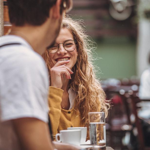 Eine lächelnde Frau mit Brille sitzt mit einem Mann in einem Café.