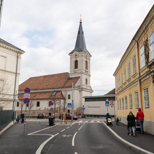Eine Kirche mit hohem Turm steht an einer Straße, umgeben von alten Gebäuden und mehreren Fußgängern.