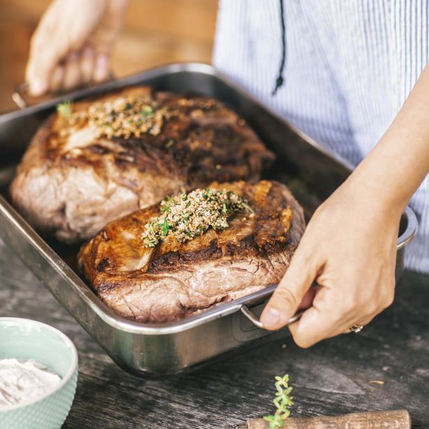 Eine Person hält ein Blech mit zwei Bratenstücken, garniert mit Kräutern und Senf.