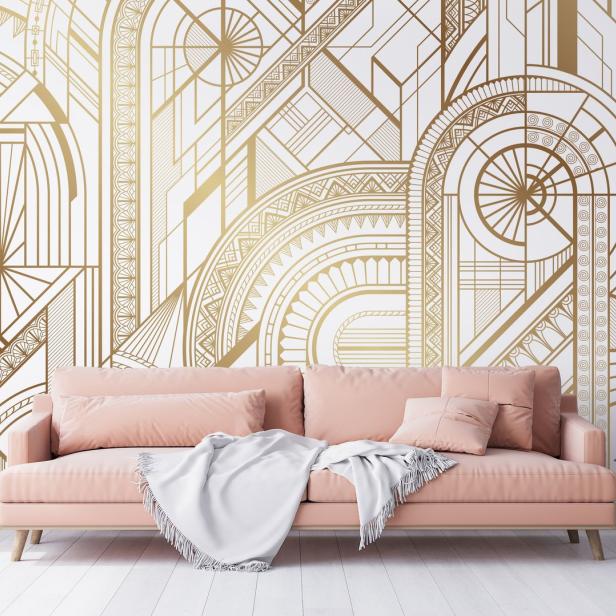 Ein rosa Sofa mit einer Decke davor steht vor einer Wand mit goldenen Art-Déco-Mustern.