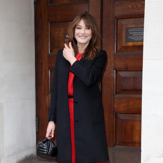 Carla Bruni trägt einen roten Hosenanzug und einen schwarzen Mantel.