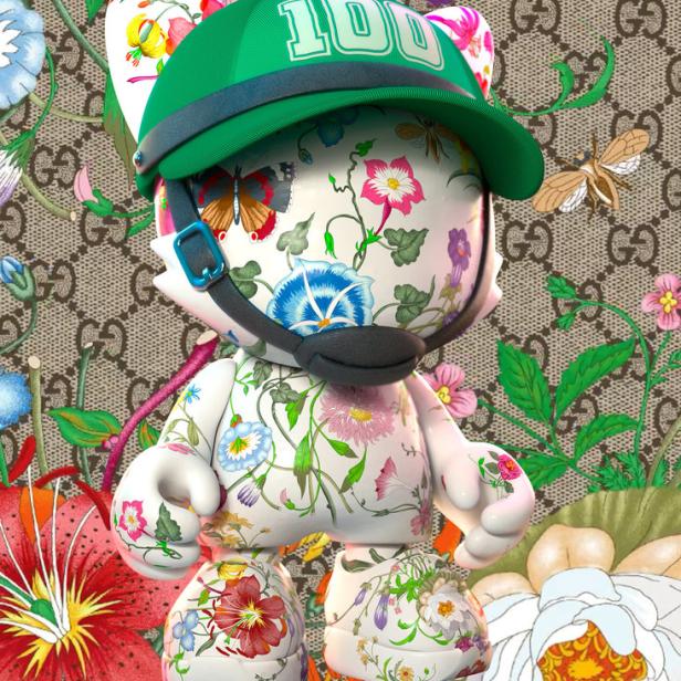 Eine stilisierte Katze mit Blumenmuster, einer grünen Kappe mit der Zahl 100 und einem Gucci-Hintergrund.