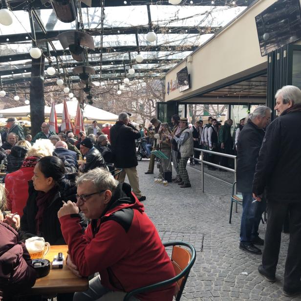 Viele Menschen sitzen und stehen in einem Biergarten, während eine Blaskapelle spielt.