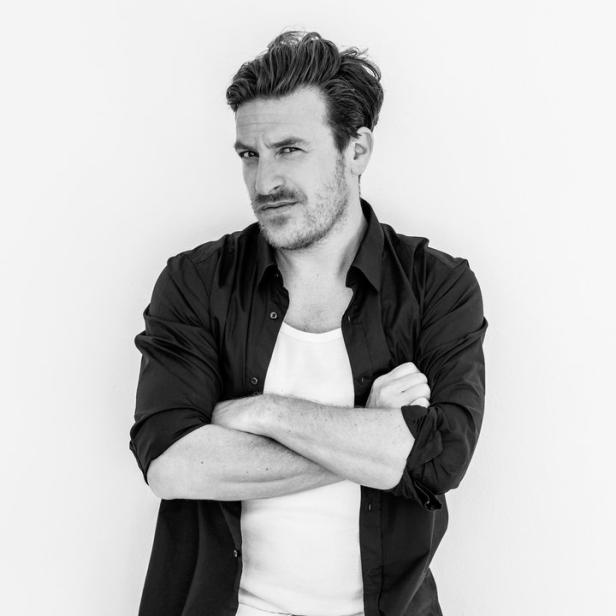 Ein Mann in Jeans und Hemd lehnt mit verschränkten Armen an einer Wand.