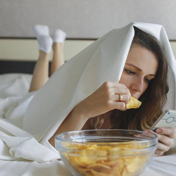Eine Frau isst Chips im Bett und schaut auf ihr Handy.