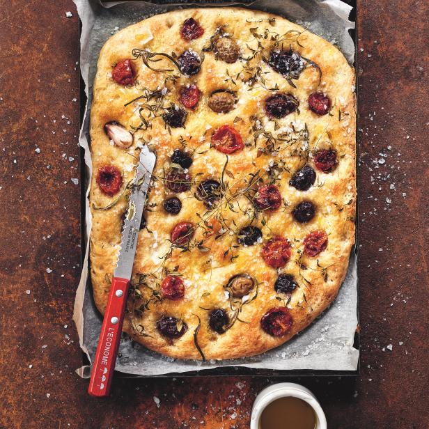 Eine Focaccia mit Tomaten, Oliven und Kräutern auf einem Backblech mit einem Messer und einer Schale Öl.